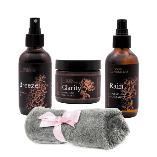 A skincare set from Good Medicine featuring three amber glass bottles (Breeze Hydrating Mist, Clarity Awakening Face Cream, Rain Replenishing Oil Cleanser) and a rolled gray microfiber towel with a pink ribbon, arranged on a white background.