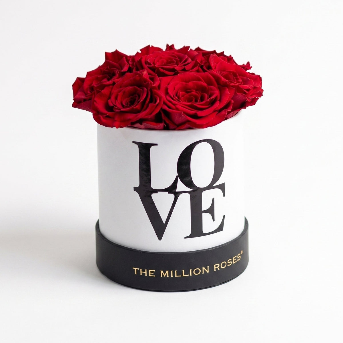 A round white luxury gift box with black lettering that reads 'LOVE' and a black base printed with 'THE MILLION ROSES', filled with a dense cluster of deep red roses on a clean white background.