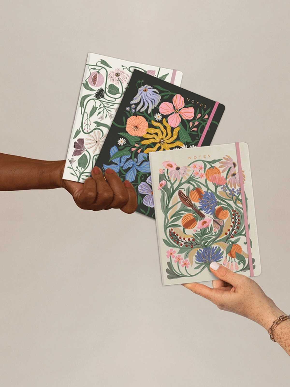Three floral notebooks held by two hands against a plain background