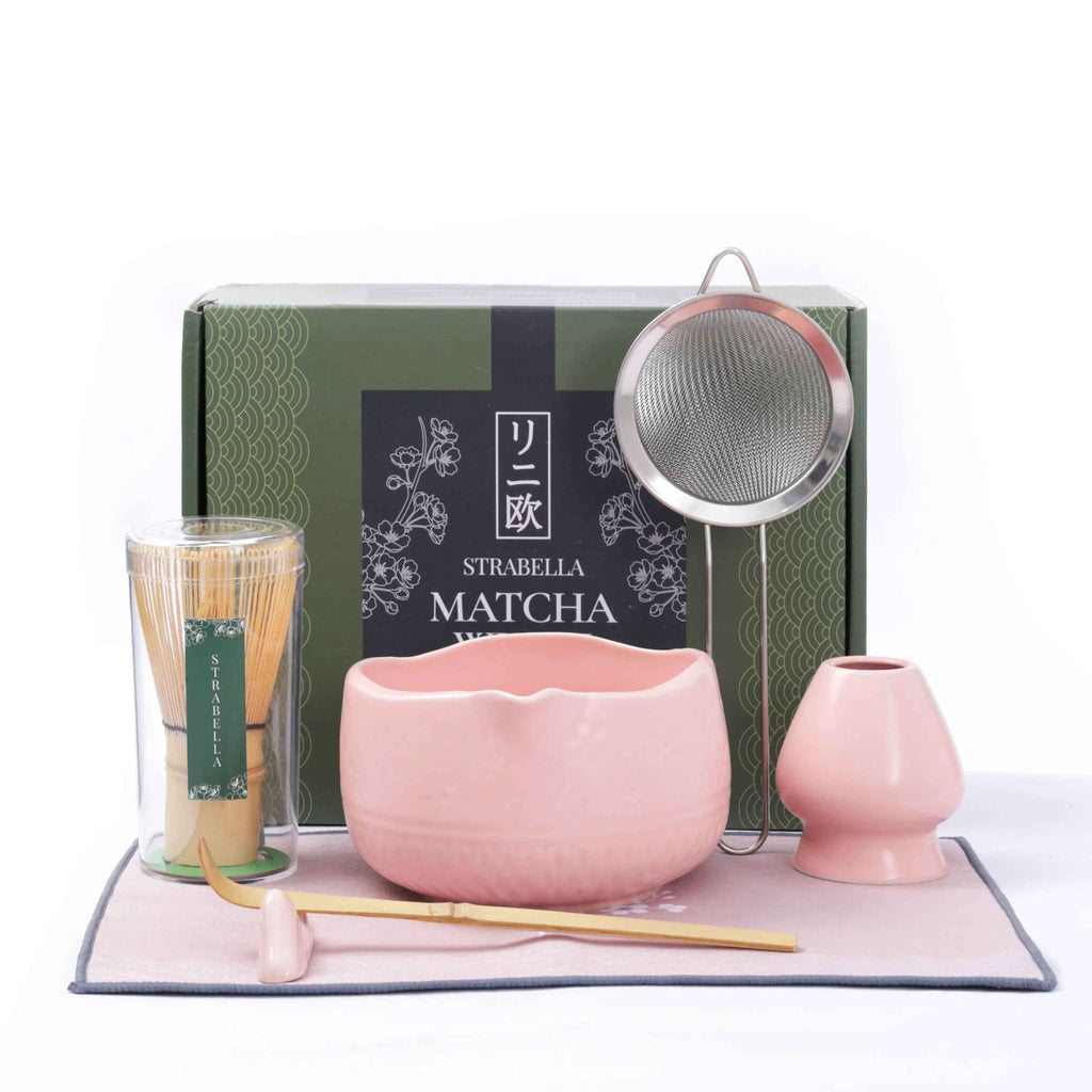 A pink matcha tea set gift set on a white background, featuring a pink ceramic bowl, matching pink teapot, a bamboo whisk in a clear container, a metal strainer, and a green Strabella Matcha box.