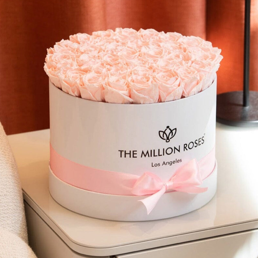 A round white gift box filled with pale pink roses, wrapped with a pink ribbon and bow. The front of the box reads 'THE MILLION ROSES Los Angeles.