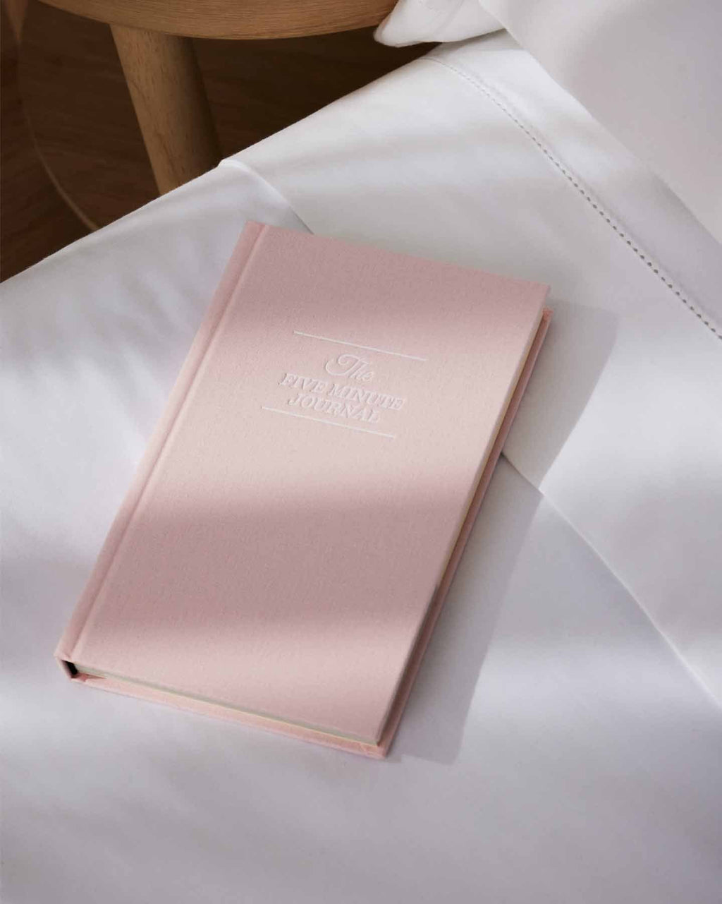 Soft pink stationery notebook labeled "The Five Minute Journal" resting on crisp white bedding next to a wooden bedside table.